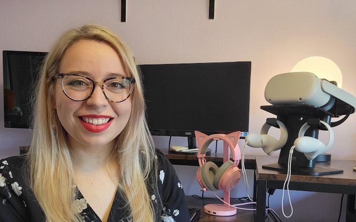 Addie in her home office, sits in front of two monitors, a VR set, and pink kitty headphones.
