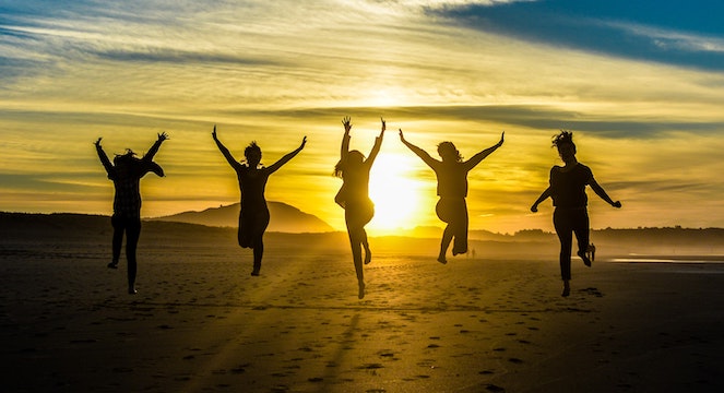 Five women taking a jumping photo at a beach at sunset