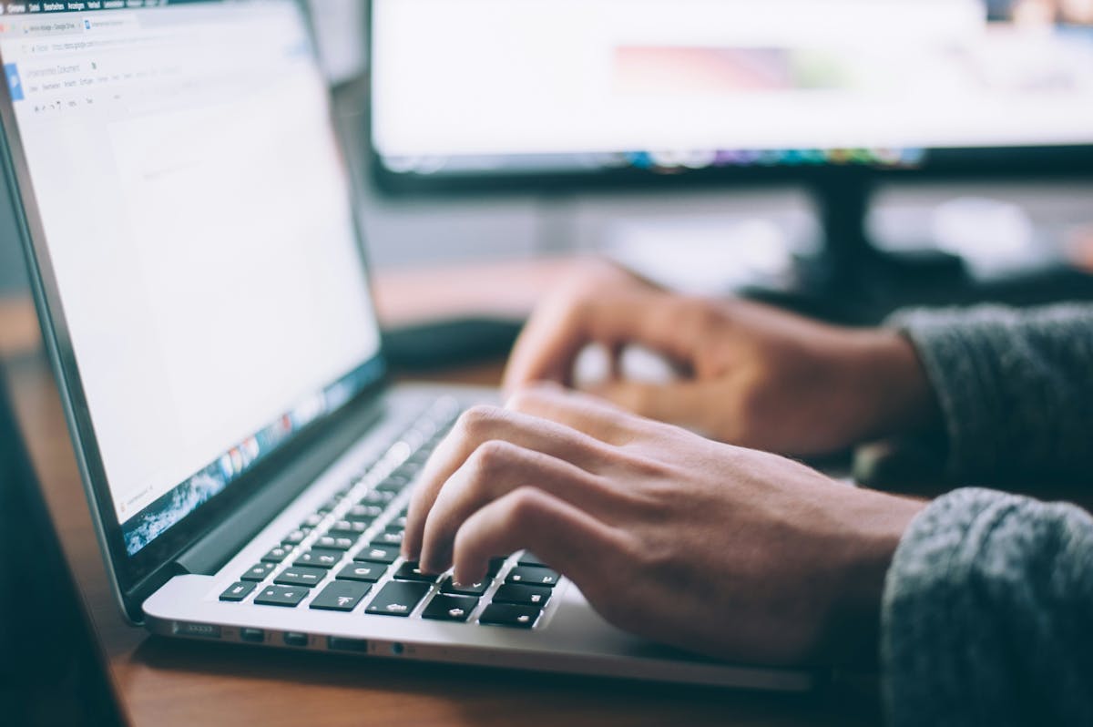 Hands typing on a MacBook laptop with Google Docs open, a second monitor visible in the blurred background on a wooden desk.