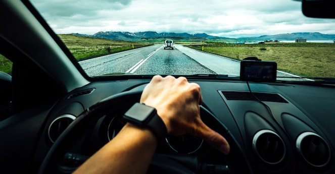 View of a hand on a car's steering wheel, driving down a country road