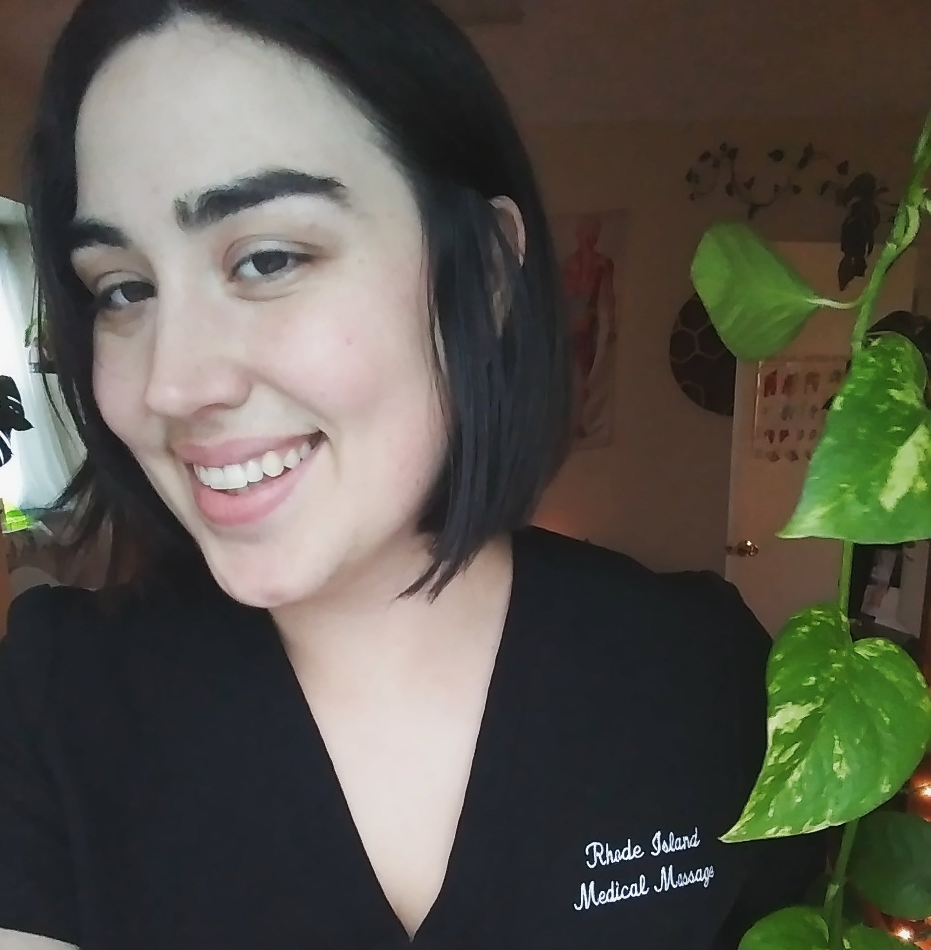 Courtney, a white woman dressed in black massage therapist scrubs, smiles.