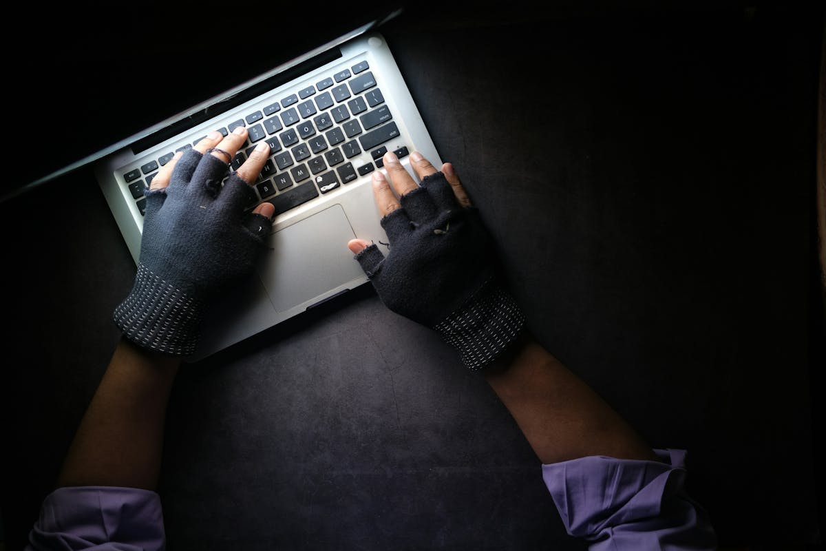 Overhead view of hands wearing black fingerless gloves typing on a silver laptop in a dark setting, evoking a hacker or cybersecurity concept.