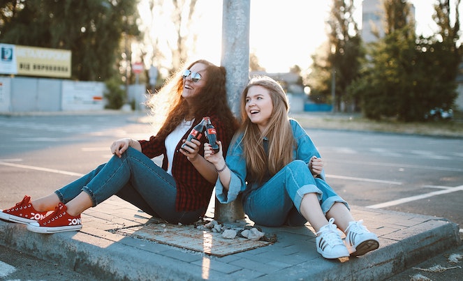 Two young girls sitting on the ground at an empty parking lot laughing together