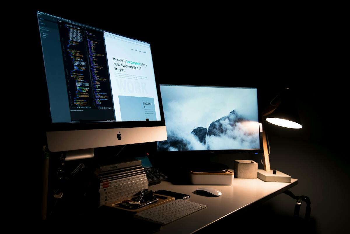 A dark desk setup with a dual monitor configuration — an iMac showing code and a portfolio website, and a second monitor displaying a misty mountain wallpaper.