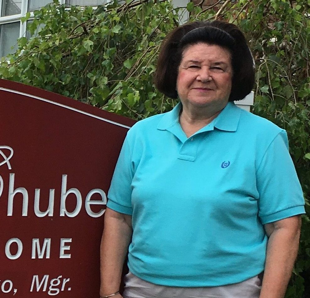 Mary Anne, a white woman in her 50s, gives a shy smile in front of the maroon funeral home sign.