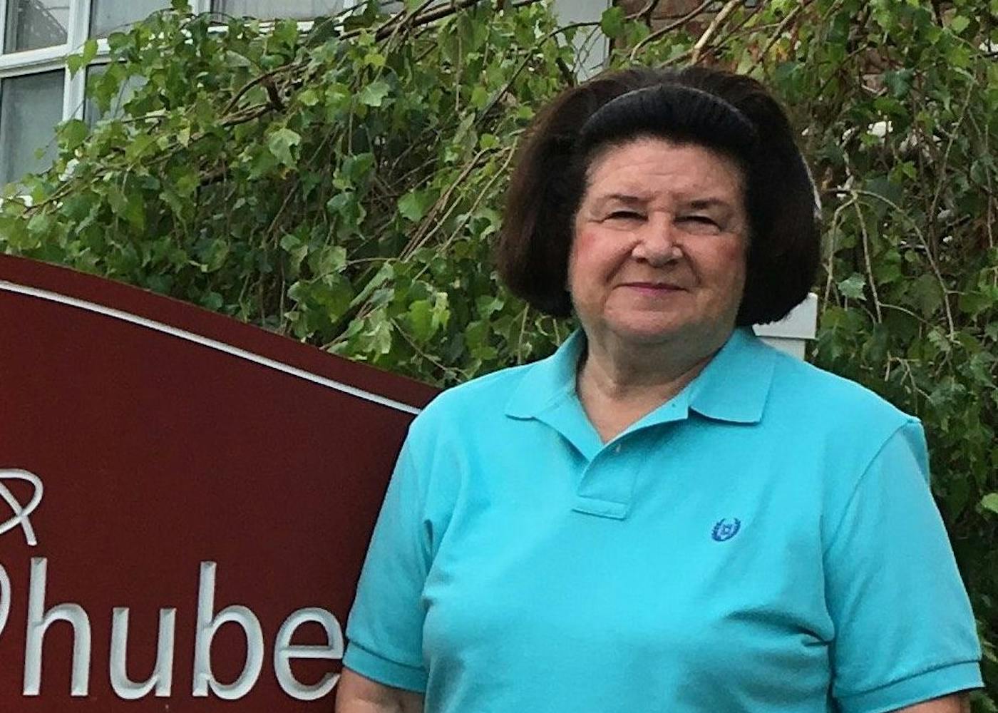 Mary Anne, a white woman in her 50s, gives a shy smile in front of the maroon funeral home sign.