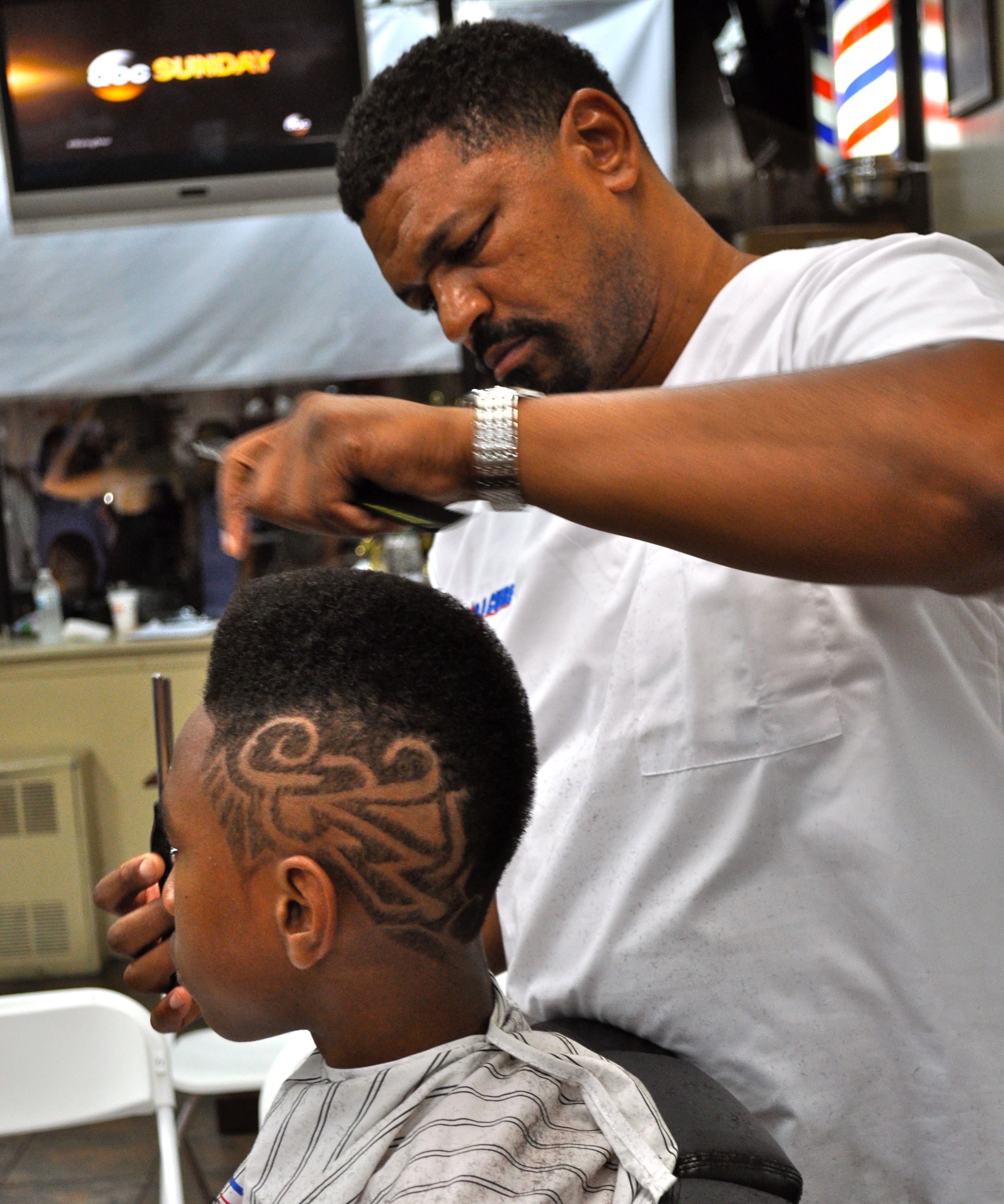 Denny, a Black man, concentrates on styling a young client's hair.