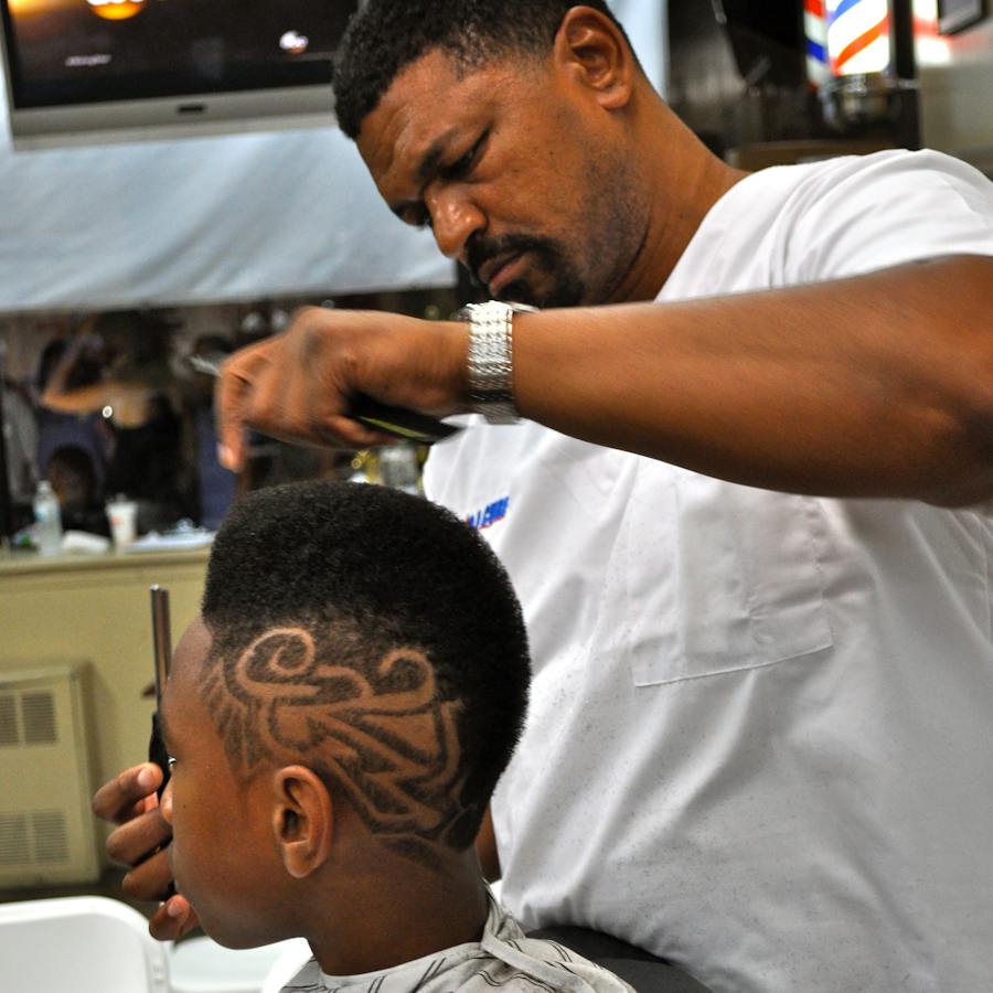 Denny, a Black man, concentrates on styling a young client's hair.