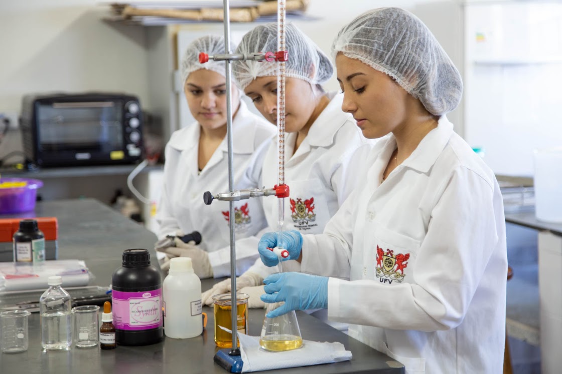Três mulheres trabalhando de touca e jaleco branco com logotipo da Universidade Federal de Viçosa utilizando instrumentos laboratoriais e analisando resultados de uma reação química. Fonte: site da reitoria da UFV