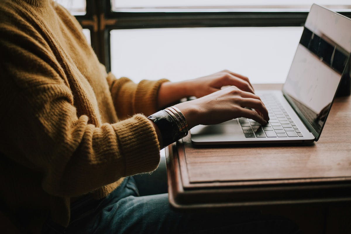 Person in a yellow sweater typing on a silver laptop at a wooden desk, wearing stacked bracelets on their wrist.