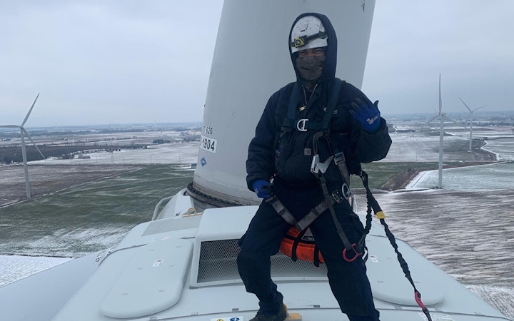 Daniel stands on top of wind turbine to make repairs in his protective hat, suit, and harness.