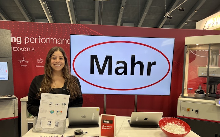 Carly, wearing a black cardigan, mans a Mahr booth at a networking event. The booth has pamphlets, candies, and Mahr pens.