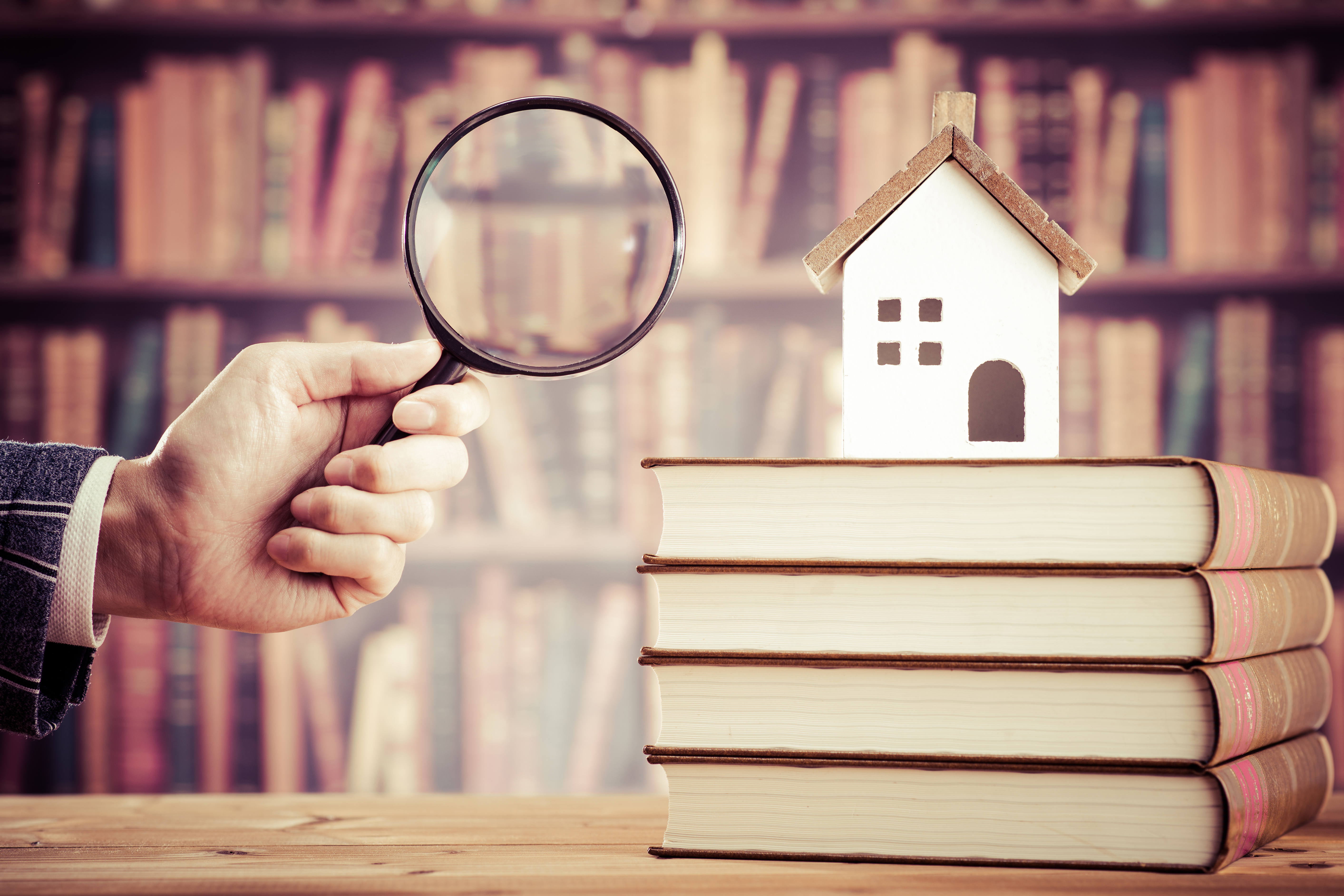 small model home on a stack of books with a hand holding a magnifying glass in the foreground