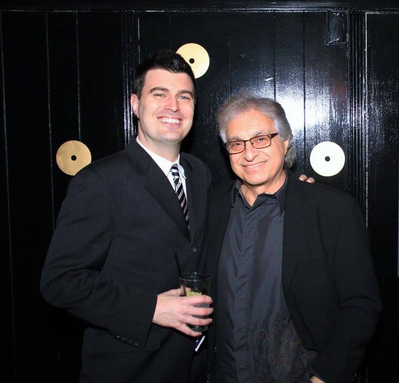 Charles, wearing a black suit and striped tie, stands next to an older man while holding a cocktail glass. Both men appear to be attending a music event.