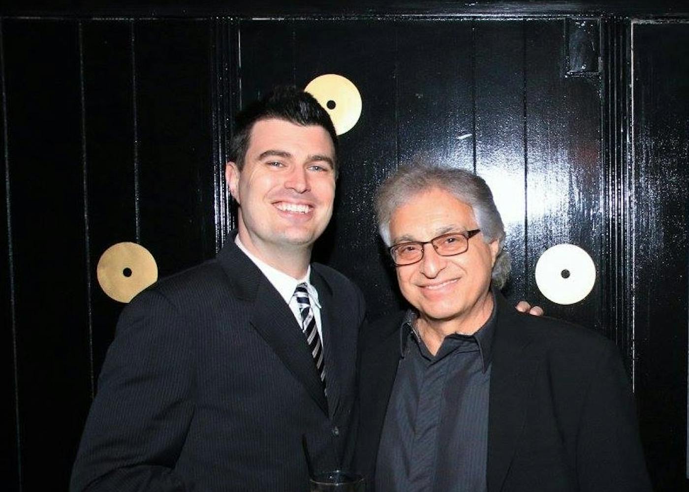 Charles, wearing a black suit and striped tie, stands next to an older man while holding a cocktail glass. Both men appear to be attending a music event.