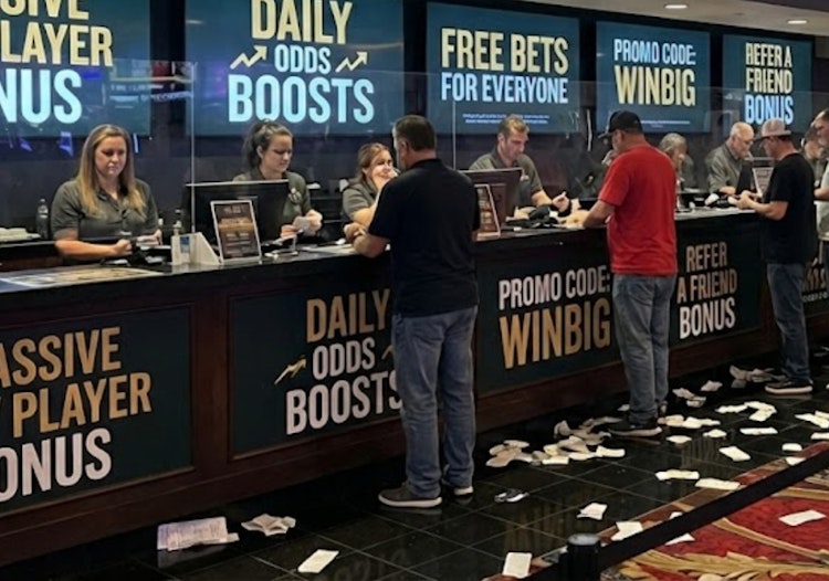 Bettors lined up at a sportsbook counter with signage advertising new player bonuses, daily odds boosts, bonus bets and promo codes, with betting slips scattered on the floor.