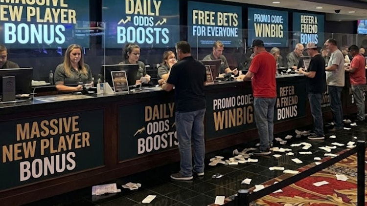 Bettors lined up at a sportsbook counter with signage advertising new player bonuses, daily odds boosts, bonus bets and promo codes, with betting slips scattered on the floor.