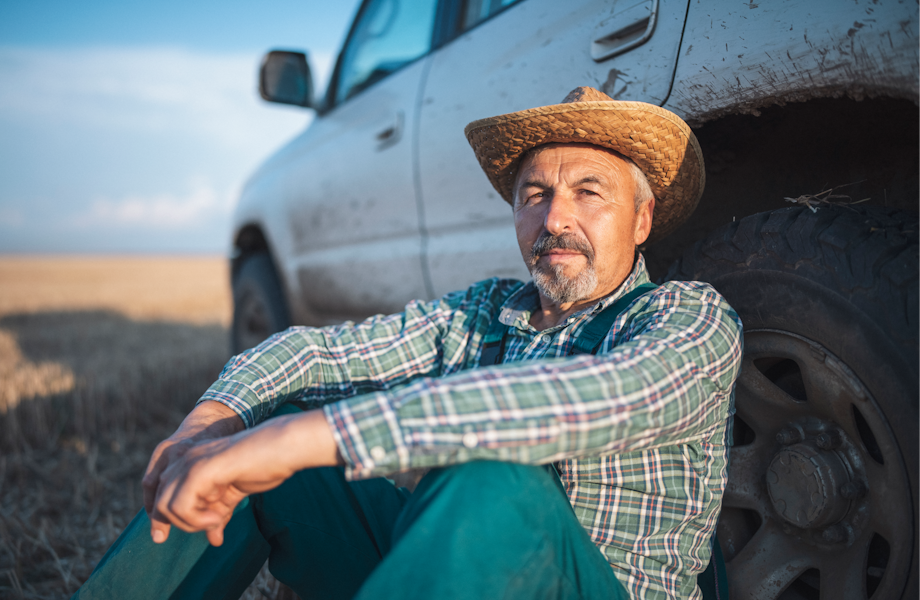 Middle age farmer leaning against a muddy truck wearing a straw hat
