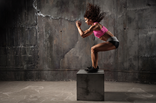Female athlete working hard and jumping up on a plyometric box with a cement background