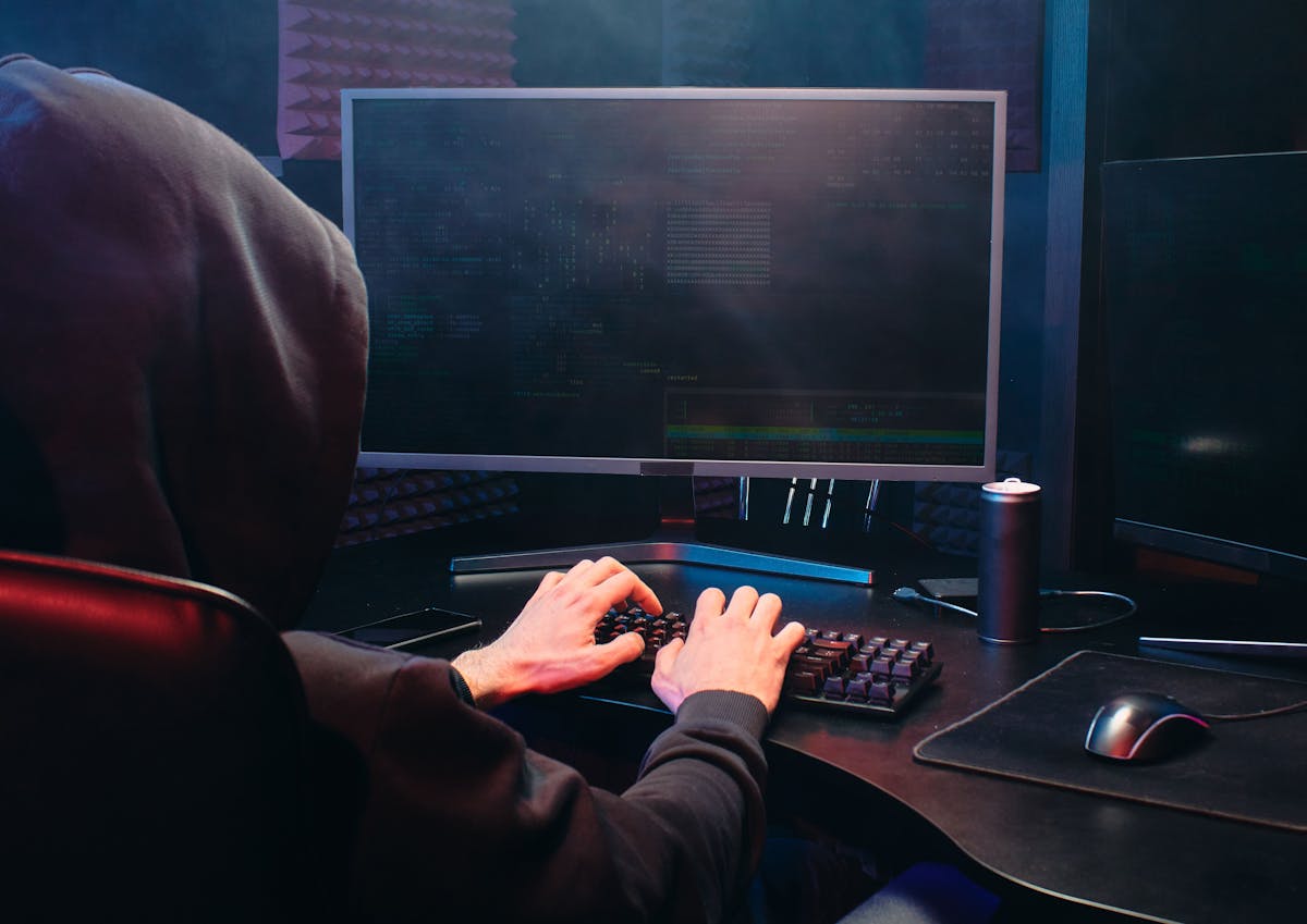 A hooded figure types on a mechanical keyboard in a dark room, with multiple monitors displaying code or terminal output, and an energy drink nearby.