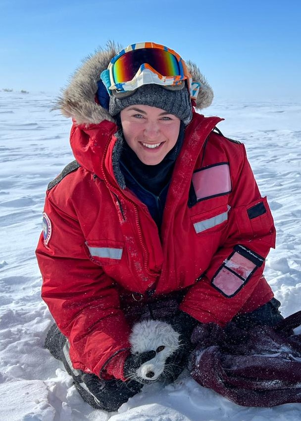 Holly wears a bright red parka, gray beanie, multicolor ski goggles, and thick warm layers in a snowy environment. She holds an animal in her grasp while kneeling on the snowy ground.