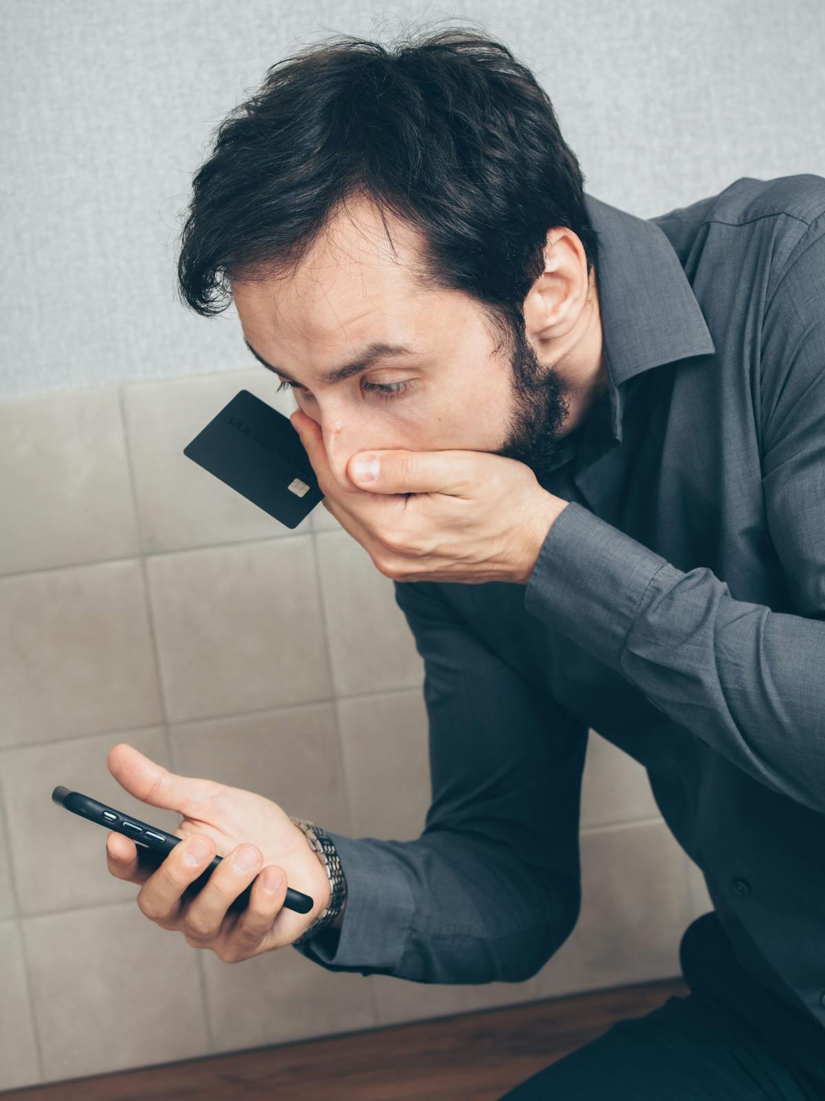 A worried man holding a credit card to his mouth while anxiously looking at his smartphone, suggesting financial stress or concern over a transaction.