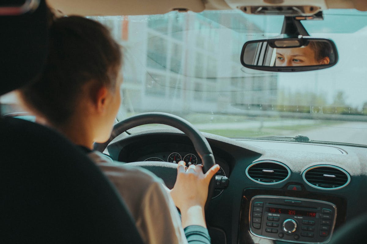 A person driving a car, viewed from behind, with their eyes visible in the rearview mirror. Dashboard and windshield visible in the foreground.