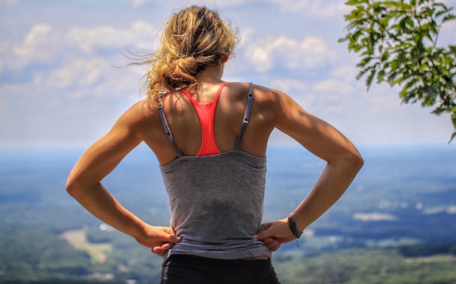 Young woman looks out at view below mountain with hands resting on her hips
