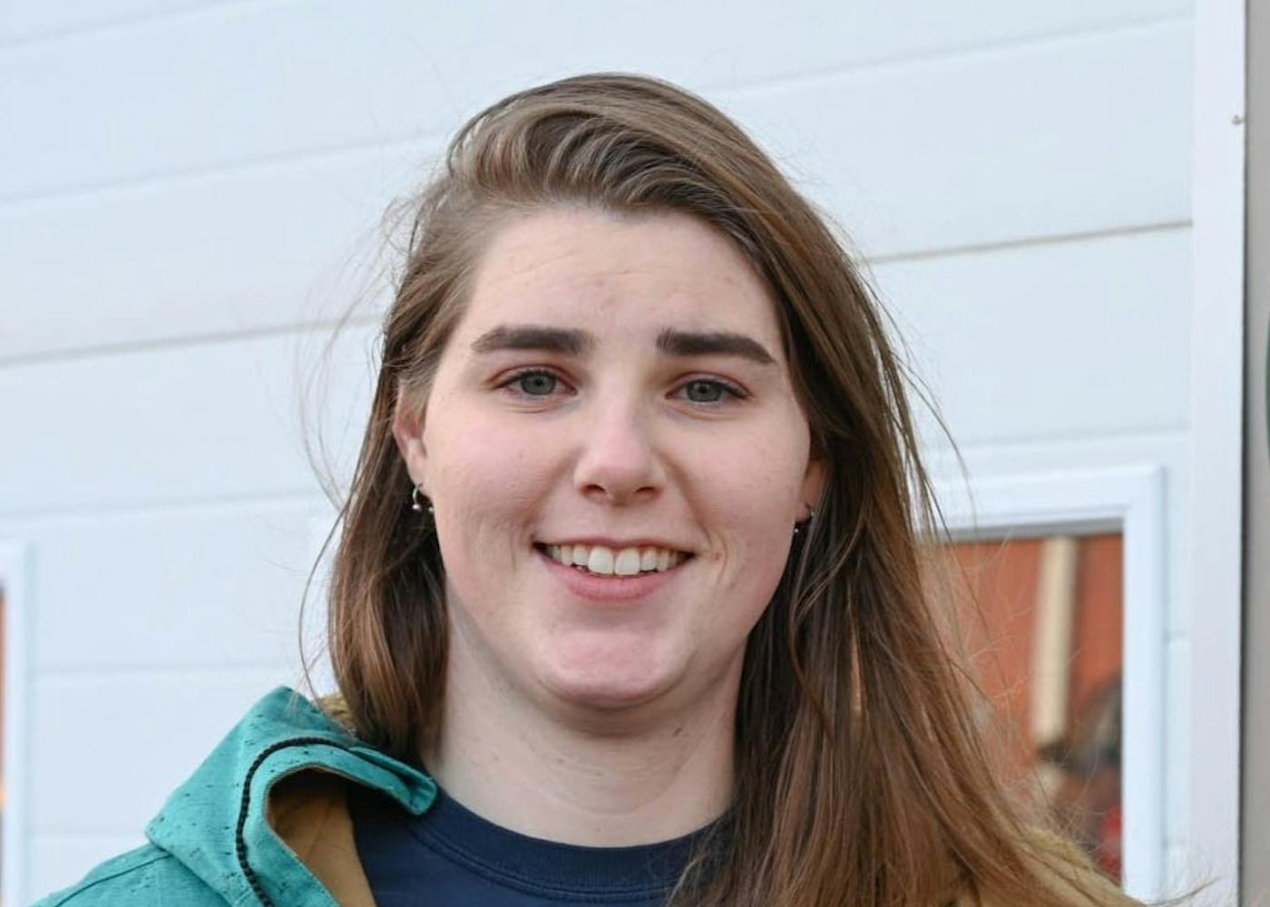 Loren, a white woman in her 20s, stands outside her work on a windy day. She is wearing a blue raincoat and dainty earrings.