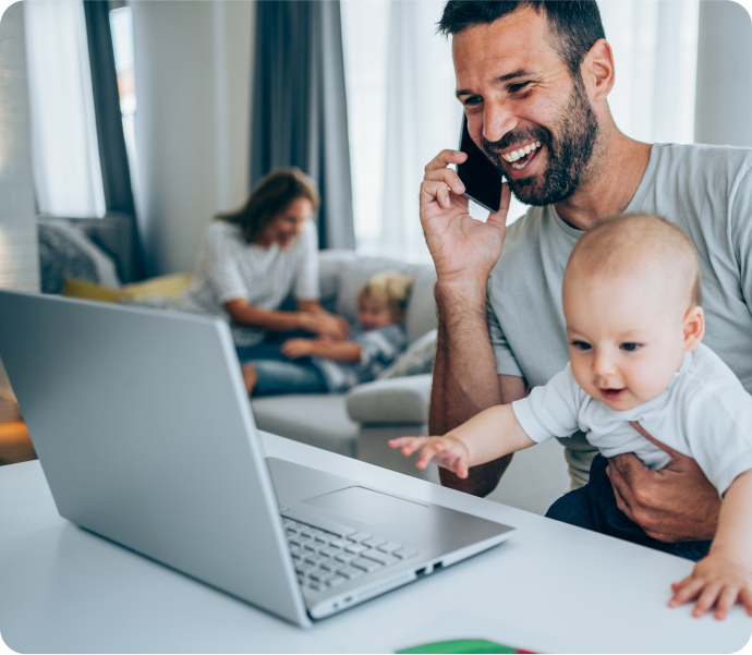 Man working on a laptop with a phone in one hand and a baby on his lap