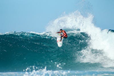 Surfer in red wetsuit rides large turquoise wave, performing aerial maneuver with white spray against clear blue sky.