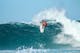 Surfer in red wetsuit rides large turquoise wave, performing aerial maneuver with white spray against clear blue sky.