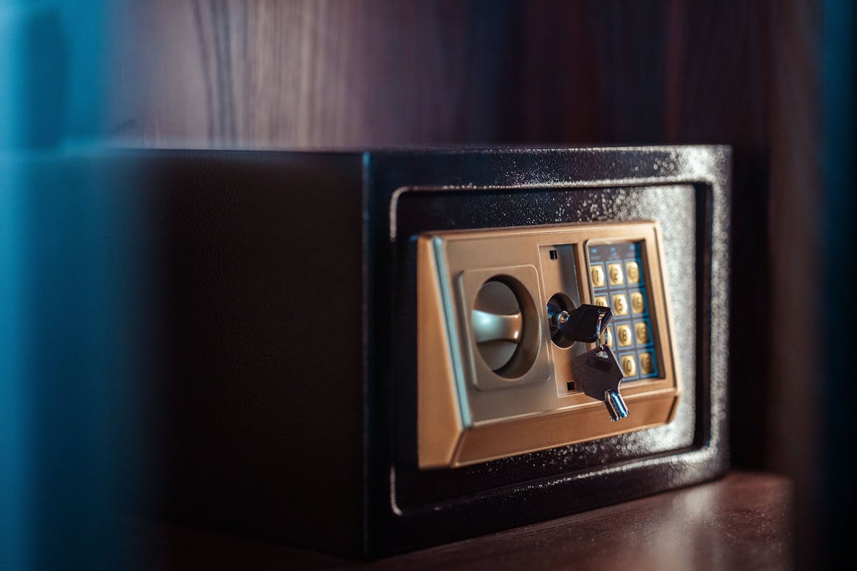 A black electronic safe with a gold keypad, key lock, and handle, mounted on a wooden shelf in a dimly lit room.