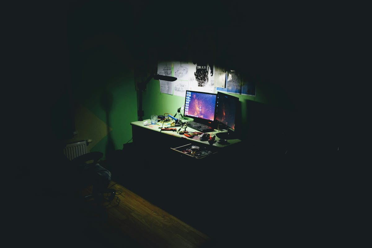 A dimly lit workspace at night featuring dual monitors with a space wallpaper, a cluttered desk with tools, a desk lamp, and papers pinned to a green wall.