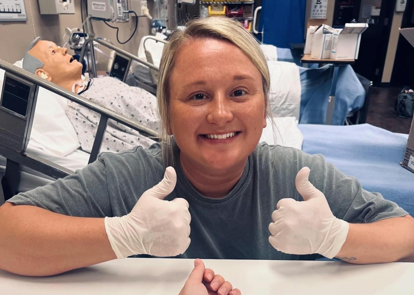 Morgan, a white woman with blond hair, wears light blue scrubs, a white long sleeve, stethoscope, and a bright smile.