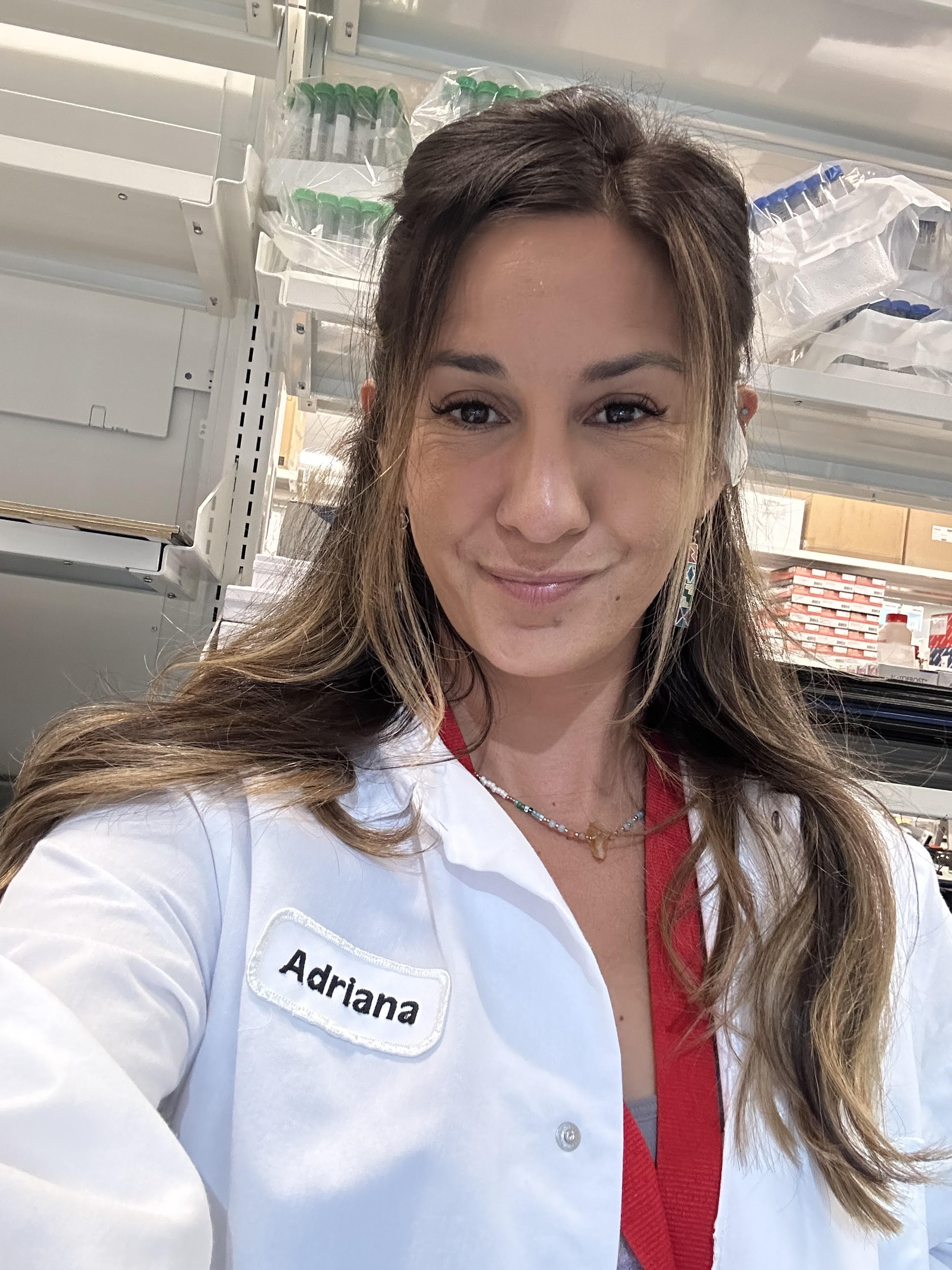 Adriana wears a white lab coat and red scrubs underneath. She is smiling in the lab.