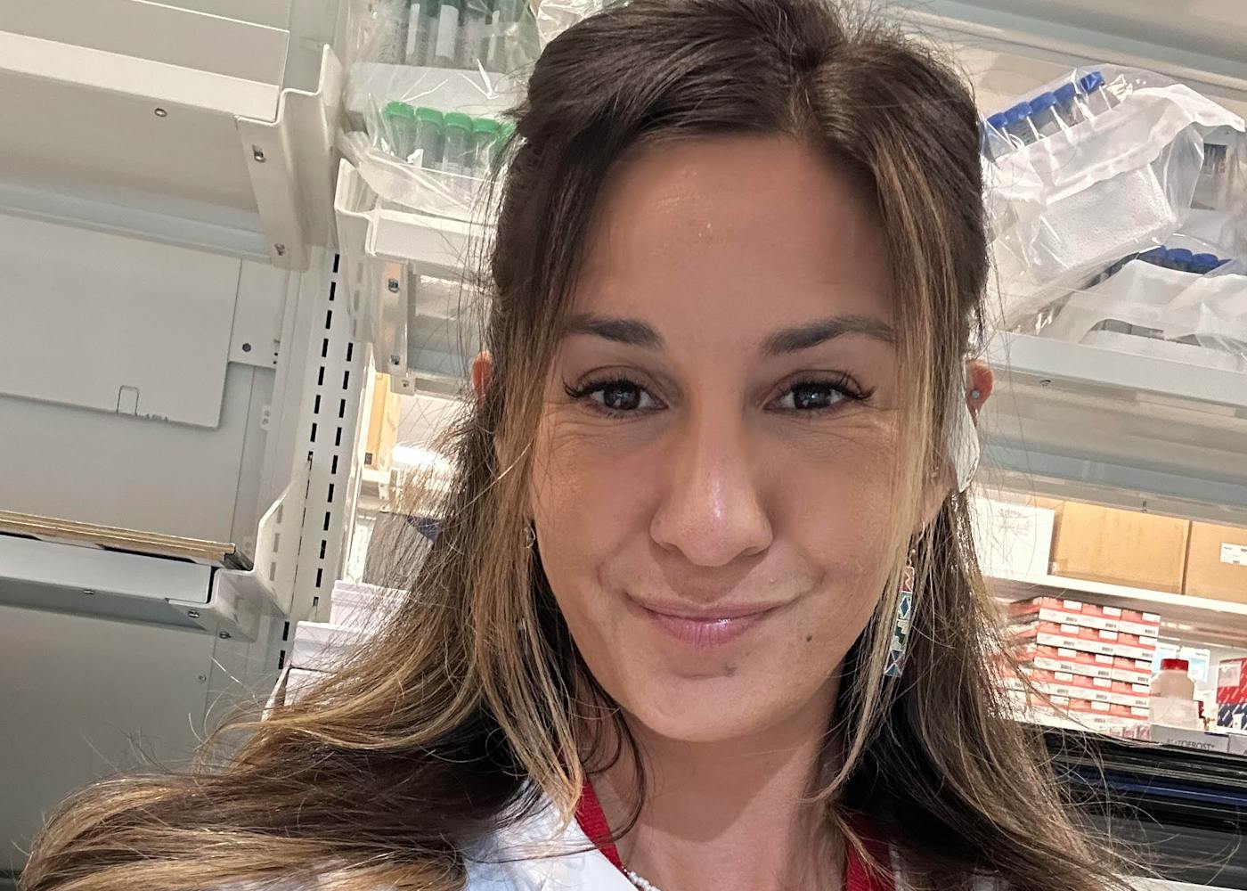 Adriana wears a white lab coat and red scrubs underneath. She is smiling in the lab.