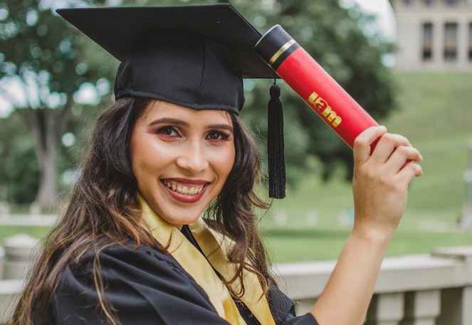 Woman wearing cap and gown smiles and poses with her graduation diploma