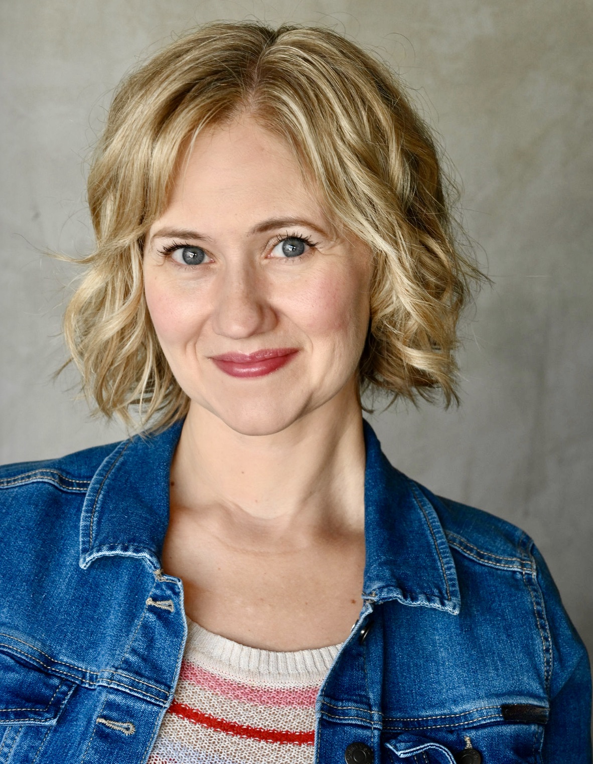 Ava, a white woman with short, blond hair, poses with a slight smile. She wears a blue denim jacket and a striped sweater underneath.