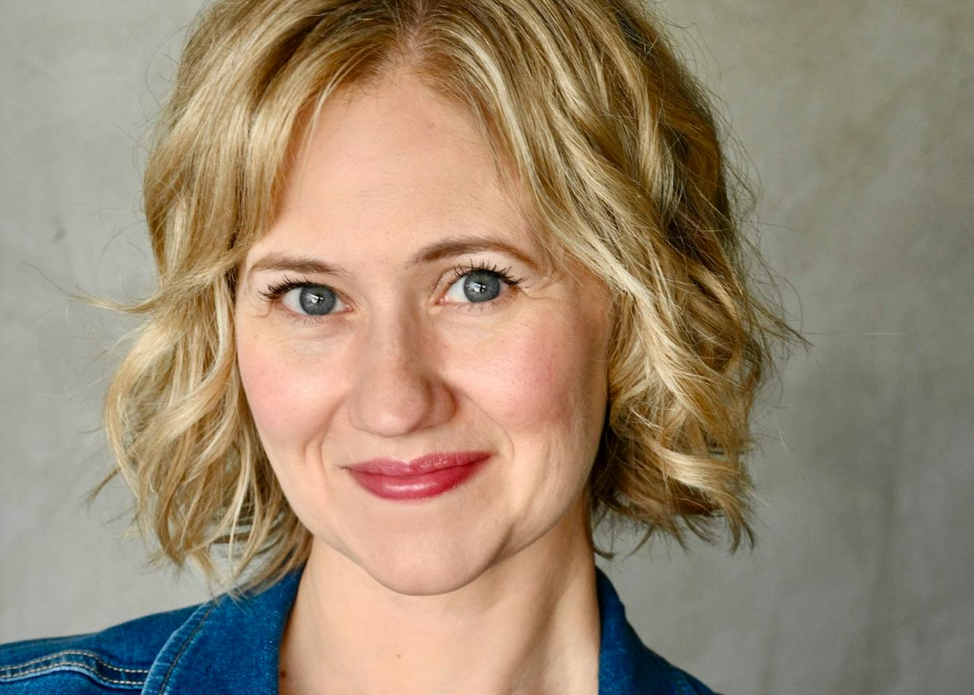 Ava, a white woman with short, blond hair, poses with a slight smile. She wears a blue denim jacket and a striped sweater underneath.