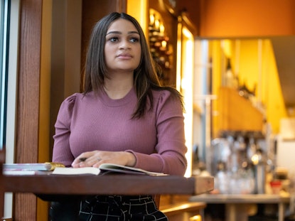 Leticia sits at a cafe table with a pen and notebook in hand as she stares into the distance.